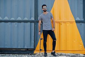 Handsome tall arabian beard man model at stripped shirt posed outdoor. Fashionable arab guy at sunglasses with skateboard against yellow painted wall. photo