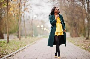 Stylish african american woman at green coat and yellow dress posed against autumn park. photo
