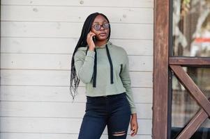 City portrait of positive young dark skinned female wearing green hoody and eyeglasses, speak on mobile phone. photo