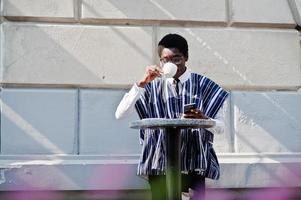 African man in traditional clothes and glasses at outdoor caffe, drink coffee and looking daily news on phone. photo