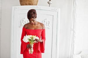 Fashion african american model in red beauty dress, sexy woman posing evening gown holding flowers in white vintage room. photo