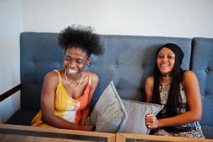 Two black african girlfriends at summer dresses sitting on the couch and fighting pillows. photo