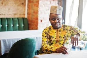 Friendly afro man in traditional clothes with cap sitting at restaurant and pours water into a glass. photo