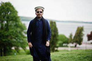 Elegante hombre árabe musulmán indio paquistaní con traje kurta dhoti, sombrero tradicional pakol y gafas de sol. foto