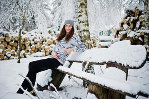 linda chica rizada con suéter y sombreros sentada en un banco en el parque forestal nevado en invierno. foto