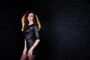 Brunette curly haired long legs girl in black leather dress posed at studio against dark brick wall. photo