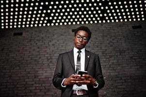 Business african american man wear on black suit and glasses at office looking on mobile phone against futuristic lights. photo