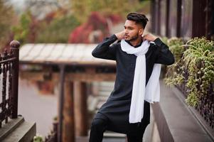 Indian stylish man in black traditional clothes with white scarf posed outdoor. photo