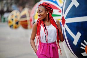 chica afroamericana de moda con pantalones rosas y rastas rojas posadas al aire libre contra huevos grandes. foto