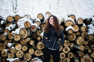 chica rizada de elegancia con chaqueta de cuero en el parque forestal nevado en invierno contra tocones. foto