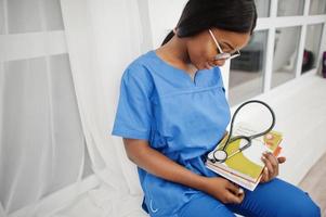retrato de una joven pediatra afroamericana feliz con uniforme azul y estetoscopio con libros en las manos. salud, medicina, especialista en medicina - concepto. foto