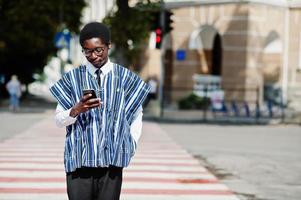 hombre africano con ropa tradicional y gafas caminando en el cruce de peatones y mirando el teléfono móvil. foto