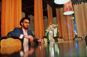 Handsome well-dressed arabian man with glass of whiskey and cigar posed at pub. photo