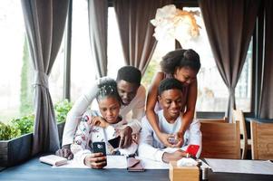 Happy african friends sitting and chatting in cafe. Group of black peoples meeting in restaurant hugs and look at their mobile phone. photo