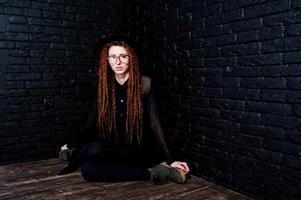 Studio shoot of girl in black with dreads, at glasses and hat on brick background. photo