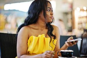 mujer afroamericana con estilo en vestido amarillo posó en el café con una taza de capuchino y teléfono móvil en las manos. foto