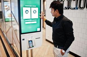 Indian man customer at store place orders and pay through self pay floor kiosk for fast food, payment terminal. Make a choise of language on screen. photo