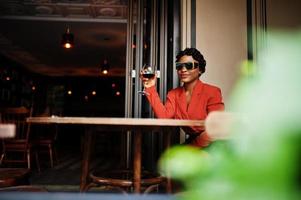 Portrait of african american woman, retro hairstyle with eyeglasses, wear orange jacket posing at restaurant with glass of wine. photo