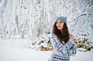 Cute curly girl in sweater and headwear at snowy forest park at winter. photo