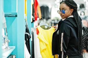 mujeres afroamericanas en chándales y gafas de sol comprando en el centro comercial de ropa deportiva contra estantes. ella elige camiseta amarilla. tema de la tienda de deportes. foto