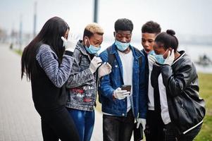 Group of african teenagers friends at park wearing medical masks protect from infections and diseases coronavirus virus quarantine, read shocked news on phone. photo
