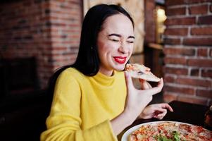 Funny brunette girl in yellow sweater eating pizza at restaurant. photo