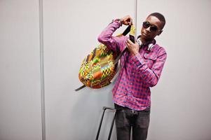 hombre afroamericano con camisa a cuadros, gafas de sol y jeans con maleta y mochila. viajero de hombre negro contra la pared gris. foto