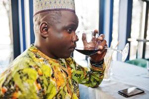 Friendly afro man in traditional yellow clothes and cap drinking red wine at restaurant. photo