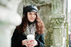 chica mexicana rizada con gorra de cuero y taza de café de plástico a mano caminando por las calles de la ciudad. foto