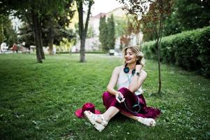 Fashionable and beautiful blonde model girl in stylish red velvet velour skirt, white blouse and hat, sitting on green grass at park with phone and earphones. photo