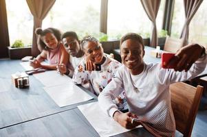 Happy african friends sitting and chatting in cafe. Group of black peoples meeting in restaurant and look at their mobile phone. photo