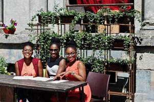 Three stylish african american womans posed at sunny summer day outdoor, sitting on table of restaurant with menu at hands. photo