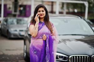 Indian hindu girl at traditional violet saree posed at street against black business suv car and speaking on mobile phone. photo