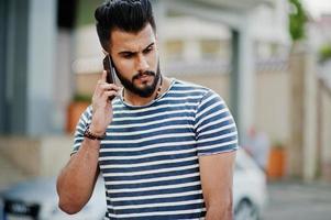 Handsome tall arabian beard man model at stripped shirt posed outdoor against car. Fashionable arab guy. Muslim businessman speaking at mobile phone. photo