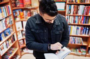 estudiante árabe alto e inteligente, vestido con chaqueta negra de jeans y anteojos, en la biblioteca con un libro en las manos. foto