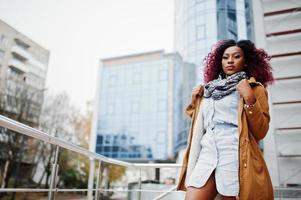 Attractive curly african american woman in brown coat posed near railings against modern multistory building. photo