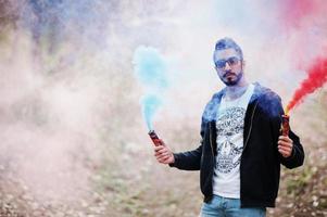 hombre árabe de estilo callejero con anteojos sostiene bengalas de mano con bomba de granada de humo rojo y azul. foto