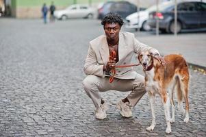 hombre afro con estilo en traje beige de la vieja escuela con perro borzoi ruso. joven africano de moda con chaqueta informal en el torso desnudo. foto