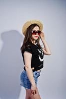 retrato de una chica atractiva en camiseta negra diciendo jajaja, pantalones cortos de mezclilla, sombrero y gafas de sol posando en el estudio. foto