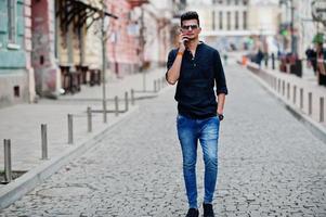 Stylish indian model man in casual clothes, black shirt and sunglasses posed outdoor at street of India and speaking at telephone. photo