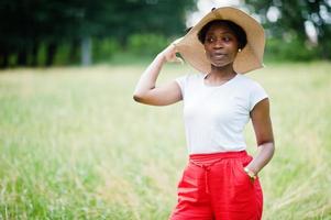 Portrait of gorgeous african american woman 20s wear in summer hat, red pants and white t-shirt, posing at green grass in park. photo