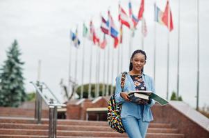 African student female posed with backpack and school items on yard of university, against flags of different countries. photo