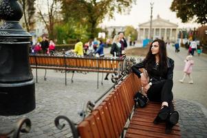 joven rizada y sexy con chaqueta de cuero sentada en un banco en la calle. foto