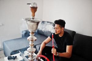 Handsome and fashionable indian man in black sitting at room, smoking hookah and working on laptop. photo