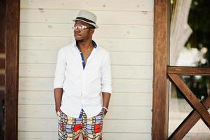 Stylish african american man in white shirt and colored pants with hat and glasses posed outdoor. Black fashionable model boy. photo