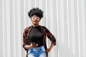 African woman with afro hair, in jeans shorts and eyeglasses posed against white steel wall with mobile phone in hand. photo