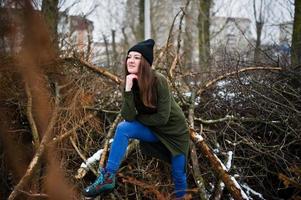 la niña usa una sudadera verde larga, jeans y sombreros negros en las ramas del pino en el día de invierno. foto