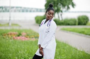 joven doctora afroamericana en bata blanca con un estetoscopio posado al aire libre. foto