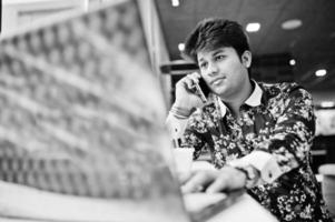 Young male indian freelancer sitting open laptop computer in fast food cafe, handsome asian man speaking phone during work on net-book in comfortable coffee shop. photo