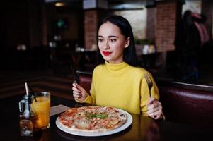 Funny brunette girl in yellow sweater eating pizza at restaurant. photo